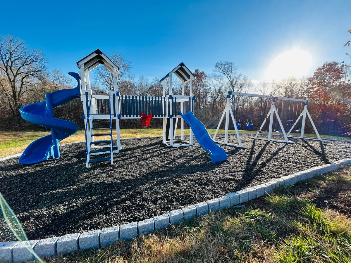 A vibrant playground setting featuring colorful slides and swings, with children playing and families gathering in the background. The area exudes energy and joy, surrounded by greenery that hints at nearby parks or recreational spaces.