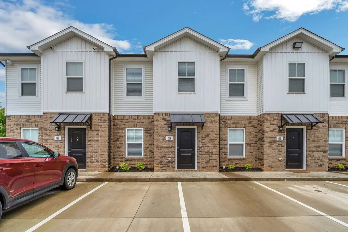 An exterior view of the apartment building featuring a classic brick facade, inviting front doors, and a convenient parking area. The scene captures neighbors gathering outside, suggesting a friendly, communal atmosphere.
