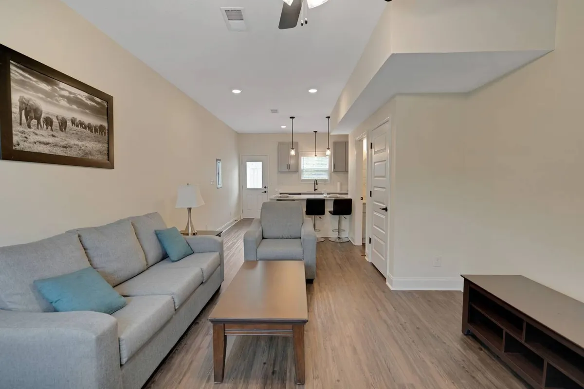 The image showcases a spacious, stylish open-plan living area featuring a comfortable sofa, modern coffee table, and a bright kitchen area in the background. Tasteful wall art adds a contemporary touch, while the layout invites relaxation, conversation, and shared meals.